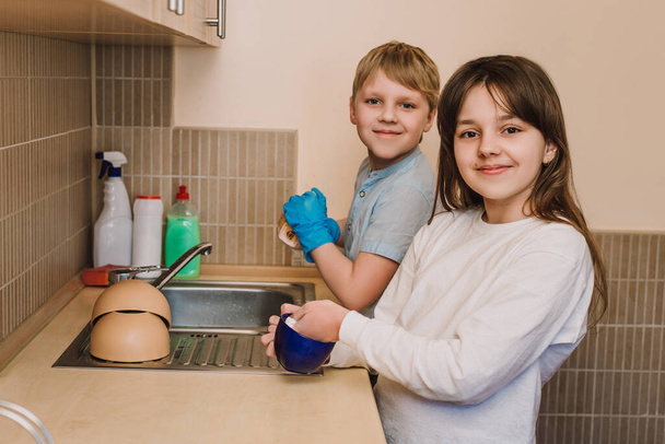 Mooi meisje en een jonge jongen die hen helpt een huishouden uit te voeren. mooi meisje en een jonge jongen in beschermende blauw rubber handschoenen wast cups in de keuken. - Foto, afbeelding