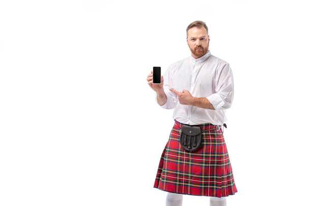 serious Scottish redhead man in red kilt pointing at smartphone with blank screen isolated on white - Foto, Imagen