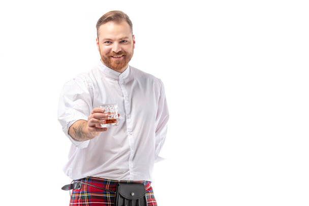 smiling Scottish redhead man in red kilt with whiskey in glass isolated on white - Photo, Image