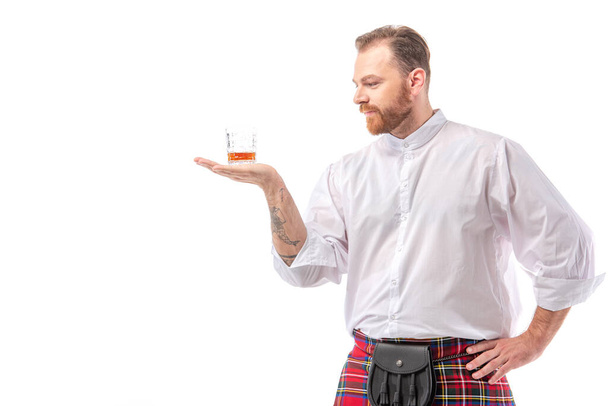 Σκοτσέζος κοκκινομάλλης σε κόκκινο κιλτ με ουίσκι σε ποτήρι στο χέρι απομονωμένος σε λευκό - Φωτογραφία, εικόνα