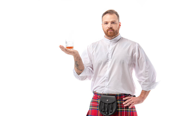 Scottish redhead man in red kilt with whiskey in glass on hand isolated on white - Photo, image