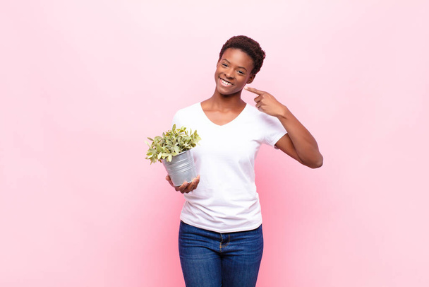 自信を持って自分の広い笑顔を指す若いかなり黒の女性,正,リラックス,植物を保持する満足態度 - 写真・画像