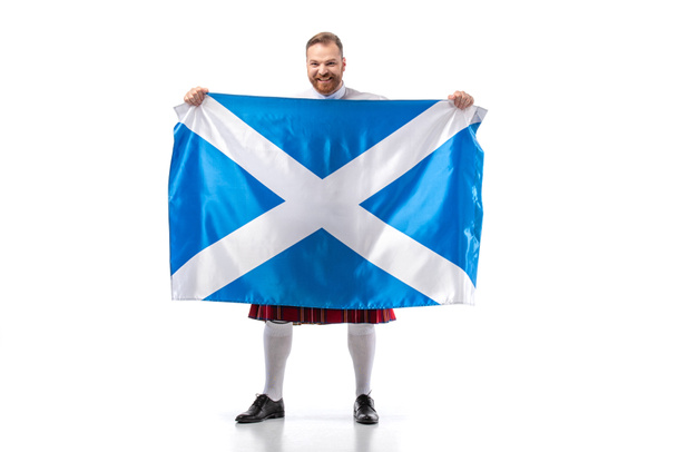 smiling Scottish redhead man in red kilt with flag of Scotland on white background - Фото, изображение