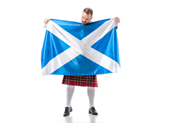 Scottish redhead man in red kilt looking at flag of Scotland on white background - Foto, Imagem