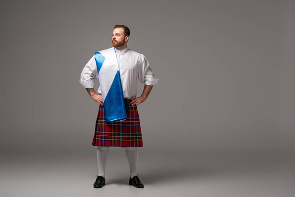 poważny szkocki rudzielec w czerwonym kilcie z flagą Szkocji i ręce na biodrach na szarym tle - Zdjęcie, obraz