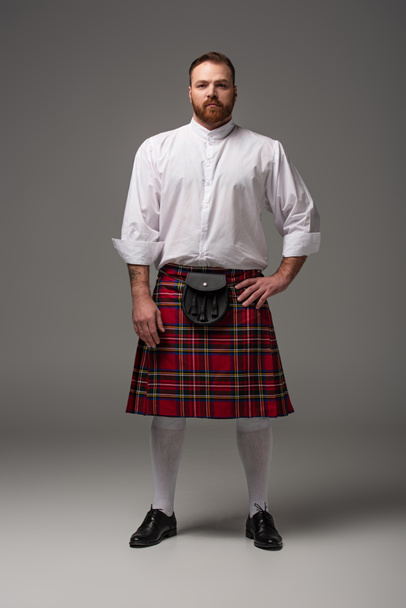 Scottish redhead man in red kilt with hand on hip on grey background - Φωτογραφία, εικόνα