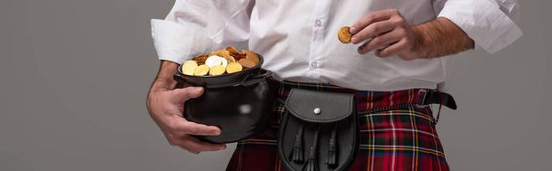 cropped view of Scottish man in red kilt holding potty with gold coins on grey background, panoramic crop - Foto, Imagen