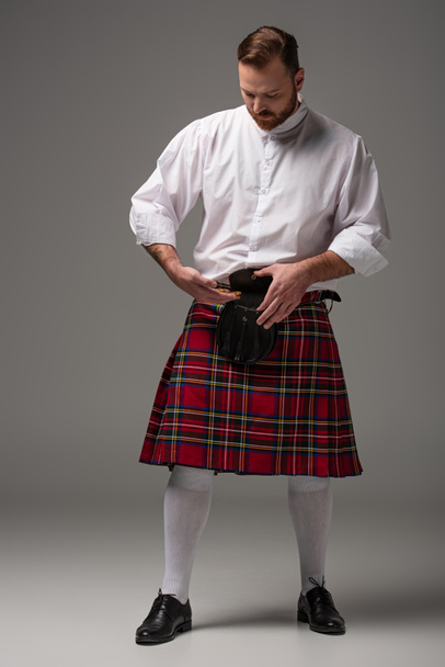 Scottish redhead man in red kilt putting gold coins in belt bag on grey background - Fotografie, Obrázek