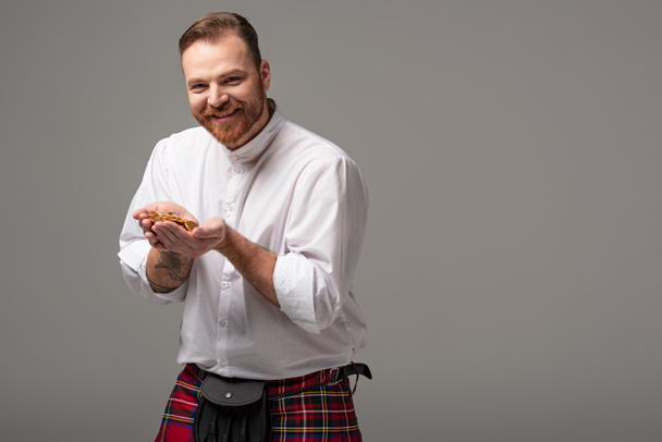 happy Scottish redhead man in red kilt with gold coins on grey background - Photo, Image