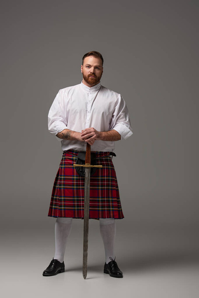 Scottish redhead man in red kilt with sword on grey background - Foto, immagini