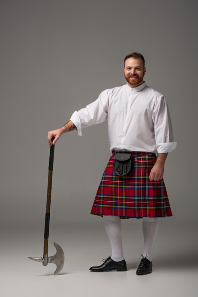 Улыбающийся шотландский рыжий мужчина в красном килте с боевым топором на сером фоне
 - Фото, изображение