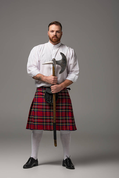 Scottish redhead man in red kilt with battle axe on grey background - Photo, Image