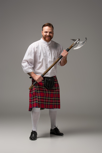 Χαμογελαστή Σκωτσέζος κοκκινομάλλης σε κόκκινο κιλτ με τσεκούρι μάχης σε γκρι φόντο - Φωτογραφία, εικόνα
