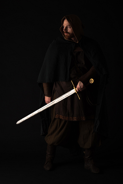 medieval Scottish man in mantel with sword in dark isolated on black - Photo, Image