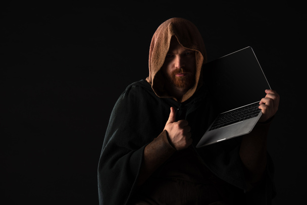 Средневековый шотландский рыжий мужчина в камине держит ноутбук и показывает большой палец в темноте изолированы на черном
 - Фото, изображение