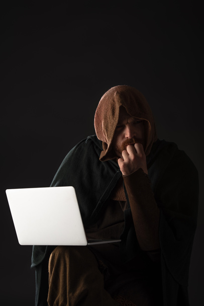 pensive middeleeuwse Schotse man in mantel met behulp van laptop in het donker geïsoleerd op zwart - Foto, afbeelding