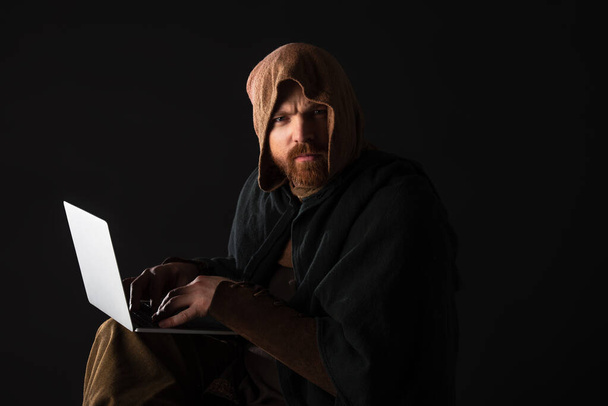 frowning medieval Scottish man in mantel using laptop in dark isolated on black - Foto, Imagen