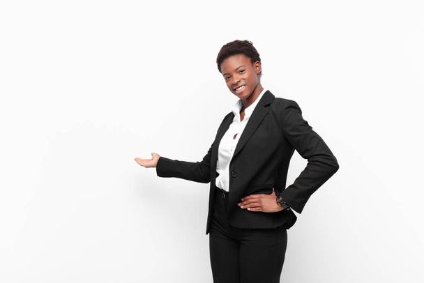 young pretty black womanfeeling happy and cheerful, smiling and welcoming you, inviting you in with a friendly gesture against white wall - Photo, Image