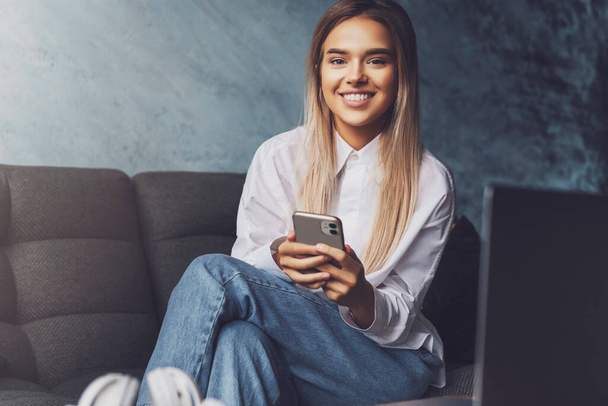 Happy smiling girl sitting on sofa, holding smartphone in hand. Young woman installing messenger app to communicate - 写真・画像