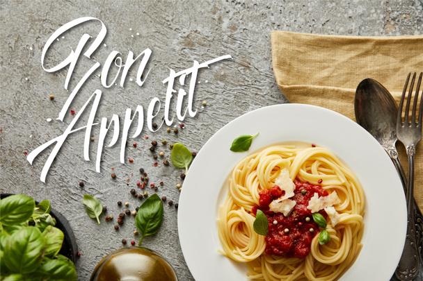 top view of delicious spaghetti with tomato sauce on plate near basil leaves and cutlery on grey textured surface with bon appetit illustration - Photo, Image
