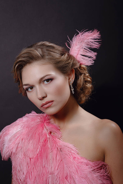 The beautiful blonde girl in a beautiful pinkostrich feather dress on black background. pleasant-looking Caucasian female looking happily in camera, emotional portrait - Zdjęcie, obraz