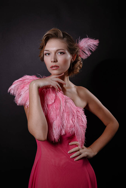 The beautiful blonde girl in a beautiful pinkostrich feather dress on black background. pleasant-looking Caucasian female looking happily in camera, emotional portrait - Фото, зображення