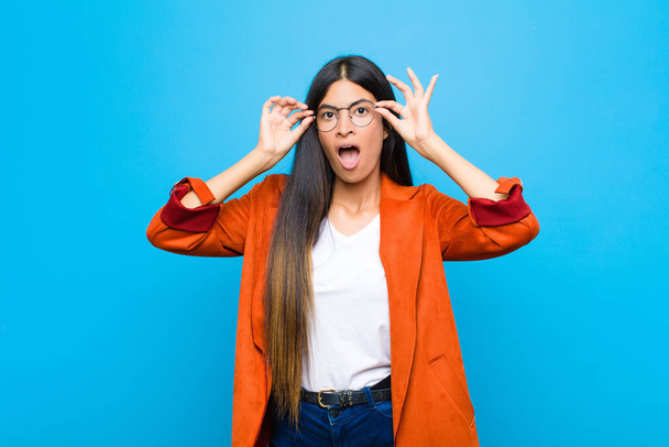 young pretty latin woman feeling shocked, amazed and surprised, holding glasses with astonished, disbelieving look against flat wall - Photo, image