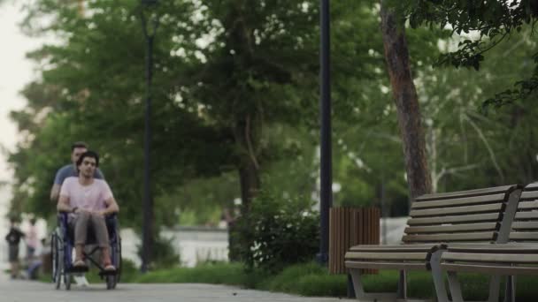 Homem deficiente feliz em uma cadeira de rodas com jovem feliz correndo no parque da cidade
 - Filmagem, Vídeo