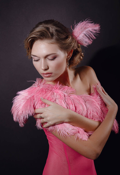 The beautiful blonde girl in a beautiful pinkostrich feather dress on black background. pleasant-looking Caucasian female looking happily in camera, emotional portrait - Foto, Imagen