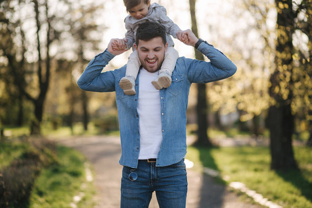 Entzückende kleine Tochter sitzt auf Papas Nacken und lacht. Junger Vater spaziert mit seiner süßen Tochter im Park - Foto, Bild