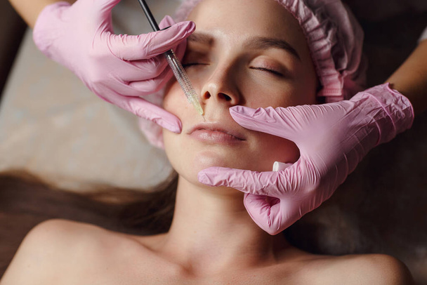 Woman on rejuvenation procedure in beauty clinic filler injection. She lying calmly at clinic. The expert beautician in pink gloves is filling female wrinkles by hyaluronic acid. Injection of beauty. - Фото, зображення