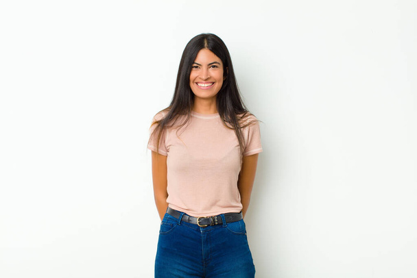 young pretty latin woman looking happy and goofy with a broad, fun, loony smile and eyes wide open against flat wall - Foto, afbeelding