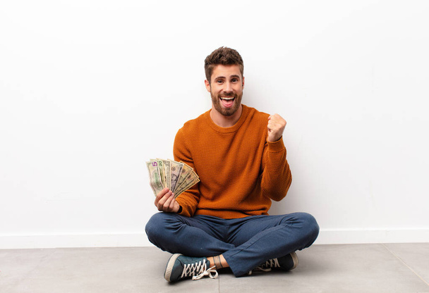 young handsome man feeling shocked, excited and happy, laughing and celebrating success, saying wow! holding dollar banknotes - Photo, image