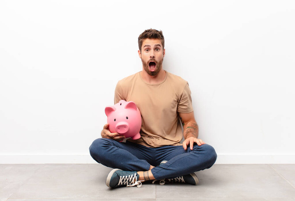 looking very shocked or surprised, staring with open mouth saying wow holding a piggy bank - Foto, Bild