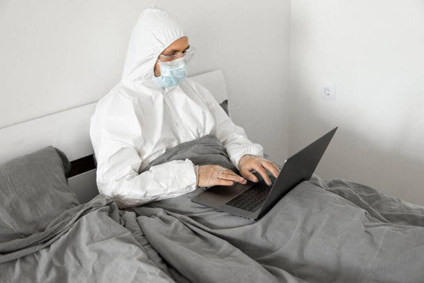Man in protective white suit and medical mask is working from home in a bed with laptop because of coronavirus epidemic. Remote work during pandemic. Stay home during COVID-19 quarantine concept. - 写真・画像