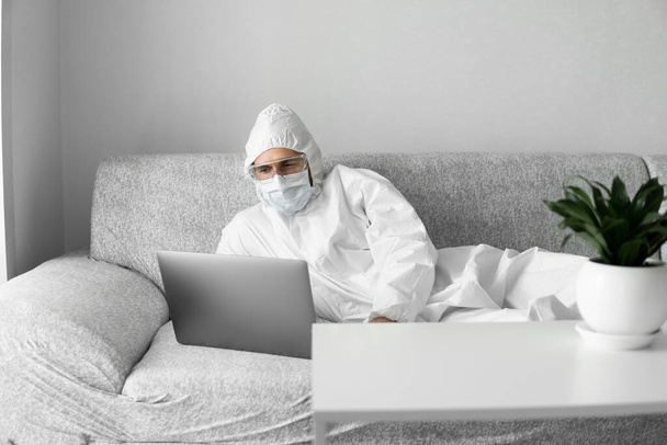 Man in protective suit and medical mask is lying on a sofa in his living room with a laptop because of coronavirus. Remote work during pandemic. Stay home during COVID-19 quarantine. Freelancer. - Фото, изображение