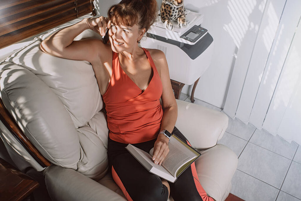 Charmante vrouw van middelbare leeftijd die praat op de smartphone en lacht, zittend met een boek zittend op een fauteuil in een woonkamer - Foto, afbeelding