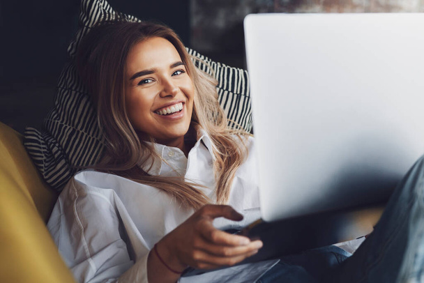 Cheerful young lady holding laptop while laying on couch. Girl relaxing after work, using internet connection - Foto, immagini