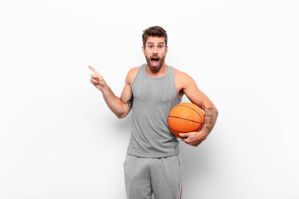 young handsome man feeling joyful and surprised, smiling with a shocked expression and pointing to the side holding a basketball ball. - Fotoğraf, Görsel
