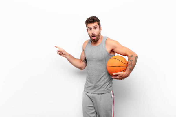 young handsome man feeling shocked and surprised, pointing to copy space on the side with amazed, open-mouthed look holding a basketball ball. - Photo, image
