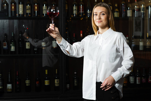 Beautiful blonde young girl sommelier holding up and looking through a glass of wine. Color and bouquet of wine. - Photo, Image