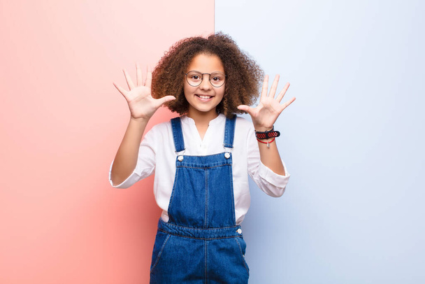 african american little girl smiling and looking friendly, showing number ten or tenth with hand forward, counting down against flat wall - Foto, afbeelding
