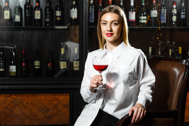 Young beautiful blonde smilling girl sommelier is holding a glass of red wine. Sommelier smelling wine bouquet. - Photo, Image