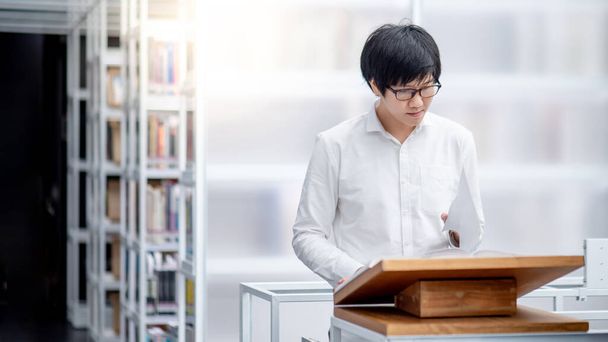 Inteligentny Azjata student uniwersytetu czyta polecaną książkę na drewnianym podium w bibliotece uczelni. Badania edukacyjne i koncepcje oppotualności stypendialnej - Zdjęcie, obraz