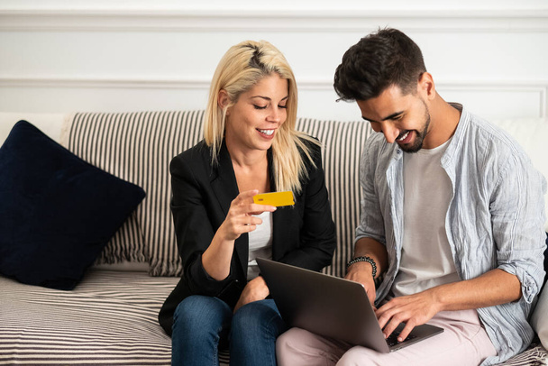 Приємна блондинка посміхається і читає кредитні картки посвідчення веселим етнічним хлопцям з ноутбуком, сидячи на дивані і роблячи онлайн покупки разом. - Фото, зображення