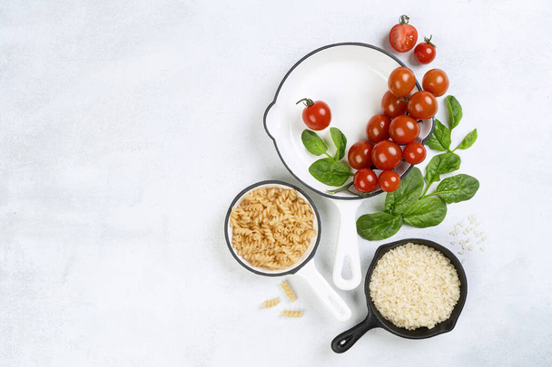Food background with vine tomatoes, spinach leaves, white rice, fusilli pasta and cast iron skillet. Background, top view, banner. Vegan or diet food concept. Composition with ingredients for cooking. - Photo, Image