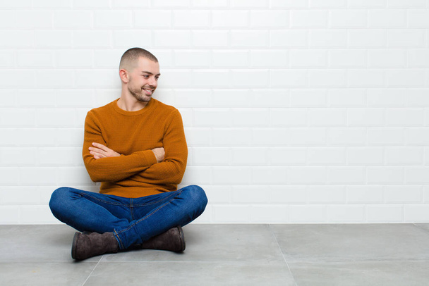 young handsome man smiling with a happy, confident expression with hand on chin, wondering and looking to the side sitting on the floor - Photo, Image