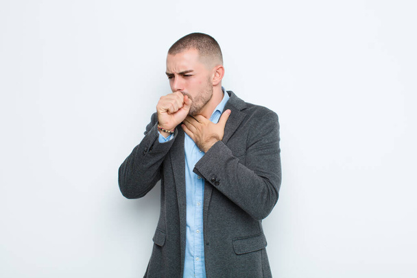 young businessman feeling ill with a sore throat and flu symptoms, coughing with mouth covered against flat wall - Foto, immagini