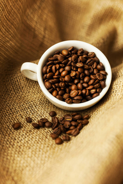 A white cup with coffee beans on burlap. A scattering of coffee beans with a cup. Coarse fabric. - Foto, imagen
