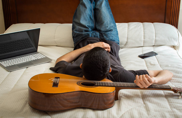 Mladý muž spí s kytarou jako polštářem. Nudný muž ležící v posteli po použití technologie a hraní na kytaru doma. - Fotografie, Obrázek